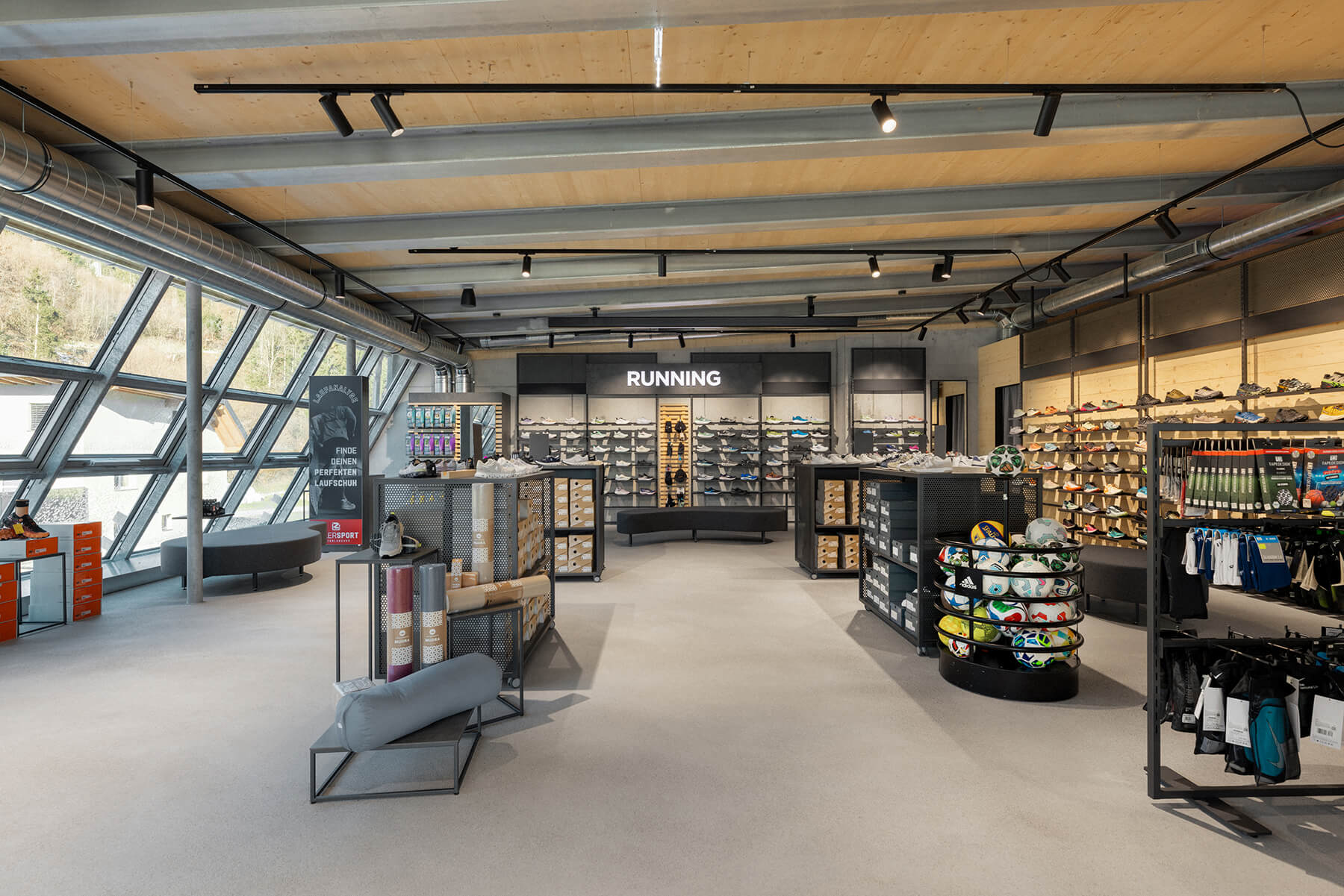 Blick durch ein Sportgeschäft lockere/durchlassende Mittelraummöbel und Wandanlagen. Keine Rückwände. Die Sicht gehr direkt auf die Sichtbetonwand oder Täferwand. Verwendung von schwarzbeschichtetem Metal