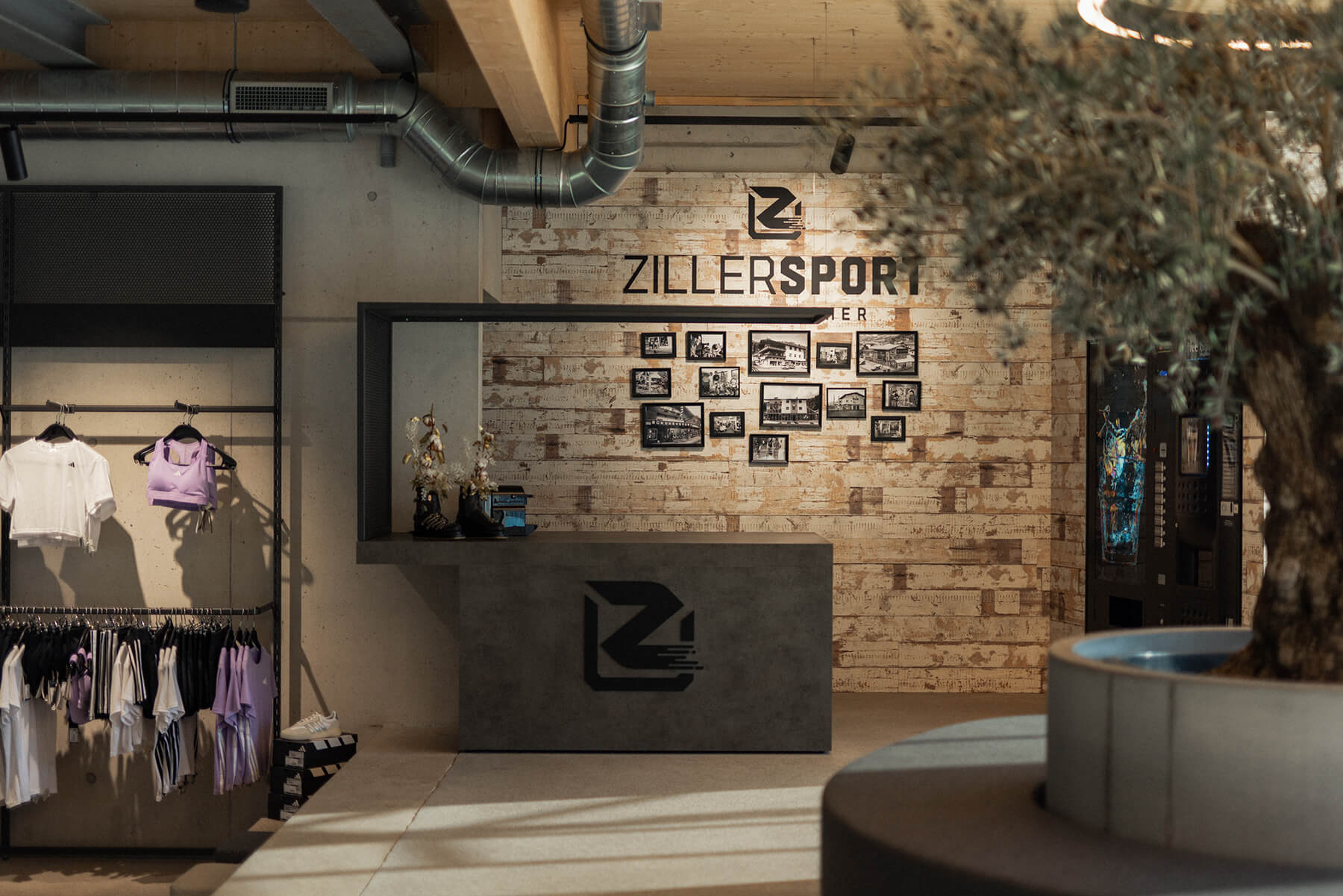 Blick auf einen Barbereich in einem Sportfachhandel. Täfer an der Wand mit Beschriftungen des Unternehmens und der Automaten zur Getränke Bedienung. Bartheke mit schwebenden Überdachten Bereich und Logo. Metall in schwarz ausgeführt, teilweise mit Lochblech. Bar in dunkler Betonoberfläche. Bodenbelag ist Estrich geschliffen.